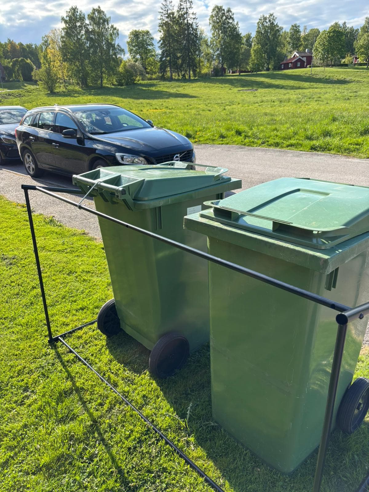 Två gröna soptunnor uppställda i Semachs öppna soptunnegarage på en gräsmatta längs en villagata
