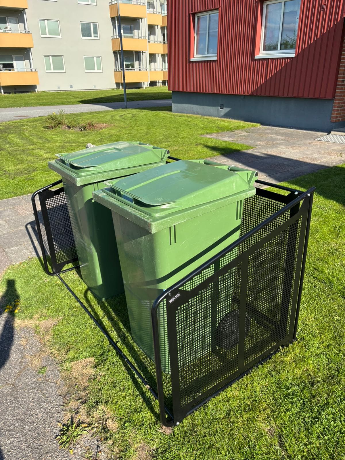 Semachs soptunnegarage med täcksidor i svartmålad metall med två gröna soptunnor utanför ett flerbostadshus