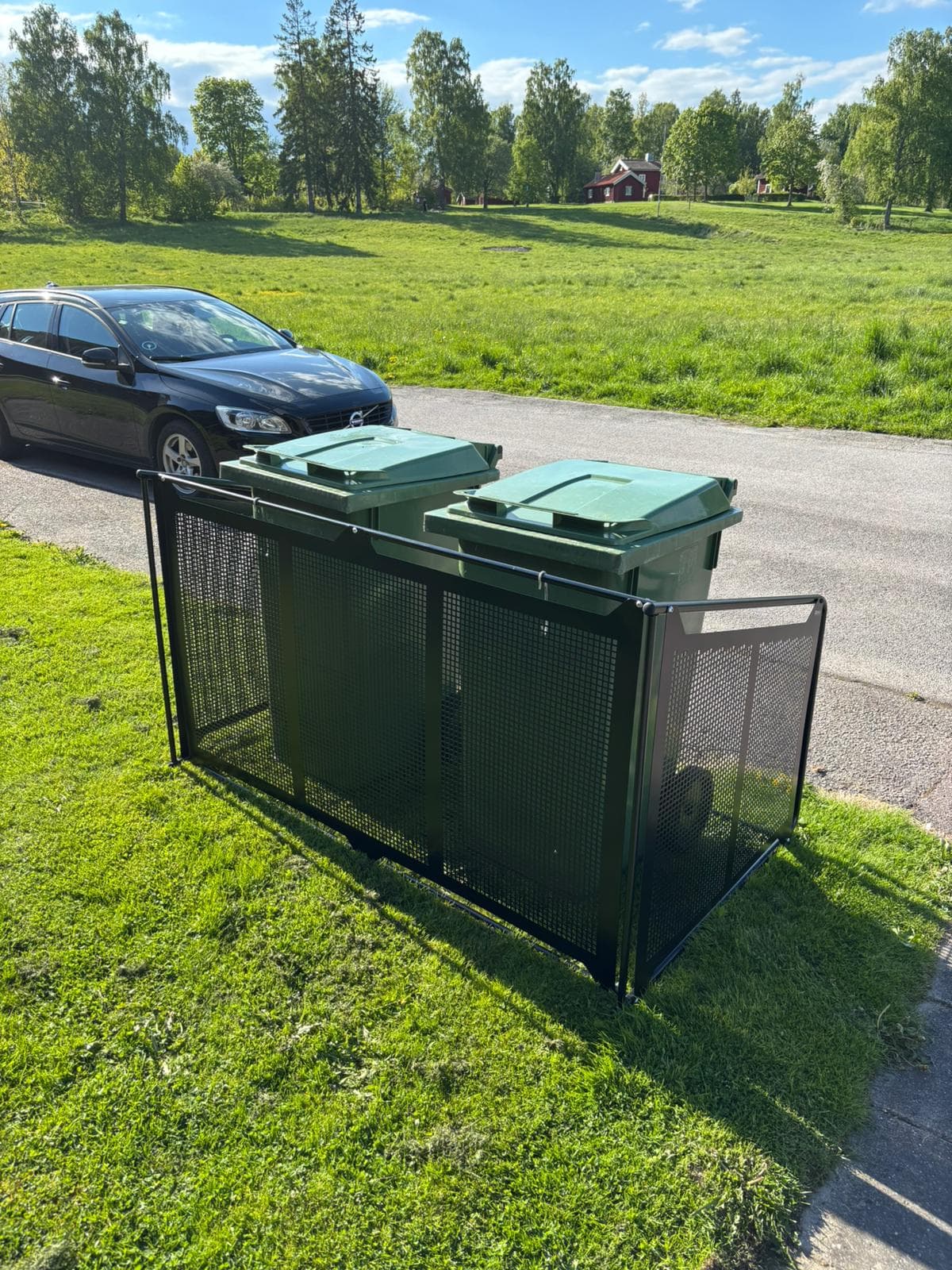Semachs soptunnegarage med täcksidor och gröna soptunnor på en gräsmatta intill en bilväg
