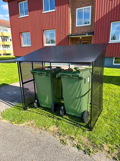 Semachs soptunnegarage med tak och täcksidor i svartmålad metall, med två gröna soptunnor uppställda utanför ett rött flerbostadshus