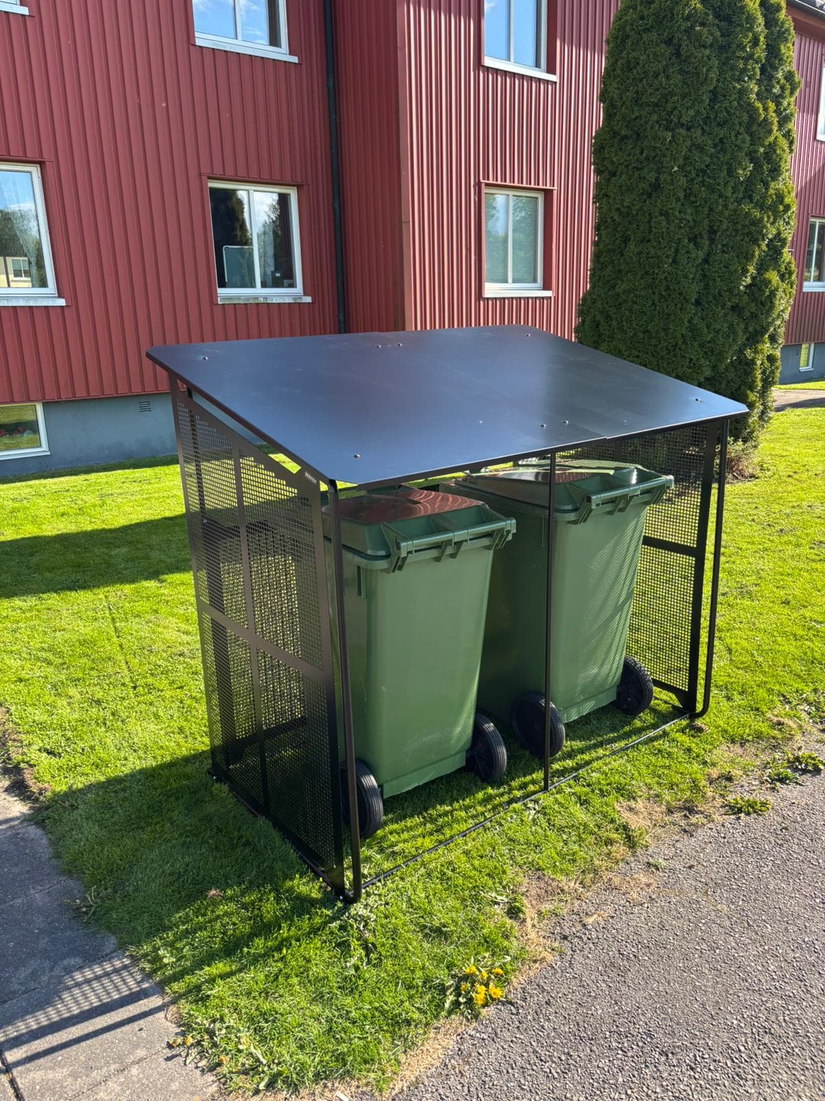 Semachs soptunnegarage med tak och täcksidor sett framifrån vid ett rött radhus, två gröna soptunnor inuti